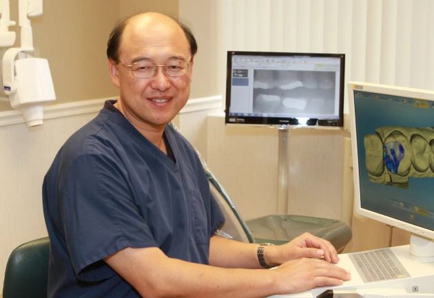 Male dentist in blue scrubs at desk with computer monitor in dental office.
